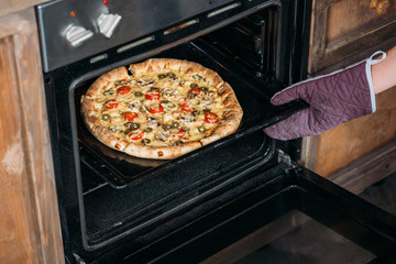 Cropped shot of young woman in kitchen glove taking pizza from oven