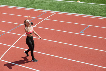 Young sporty woman athlete in sportswear drinking water or protein cocktail from shake on stadium track, workout concept
