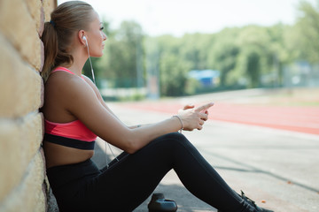 Young sporty woman athlete in sportswear sitting, listen music with headphones and relaxing on stadium track after hard workout
