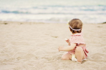 Cute baby girl with a dress on polka dots sitting on the sand on seashore and looks out to the sea