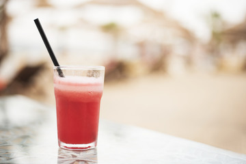 Red cocktail, watermelon fresh on a table, close up. On the background blurred umbrellas on the beach. Sun, haze, glare