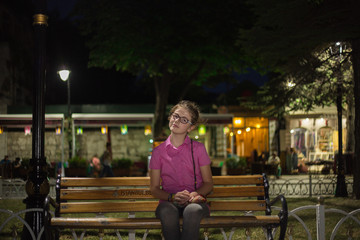 A young girl sitting on a bench in the park, eve, background lights blurred