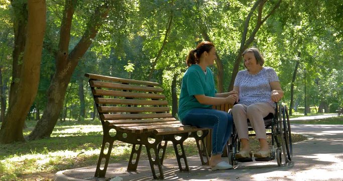 Caregiver talking to disabled senior woman in wheelchair outdoors in summer park. 4k