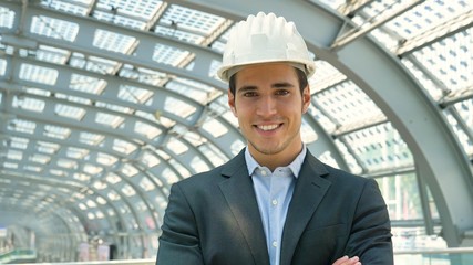 A young handsome, engineer, foreman, architect (builder) looks at the construction of the structure, at the station, wearing a white protective helmet, smiling in a suit. Concept: good work, project.