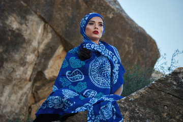 Gorgeous young long hair brunette beauty with slanted head wearing oriental colorful scarf Sits on rocks on a mountain background