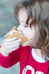 Young little girl is eating a s'more made from graham crackers, roasted marshmallows and chocolate. Her mouth is messy and she is taking a toothy bite of the s'more.