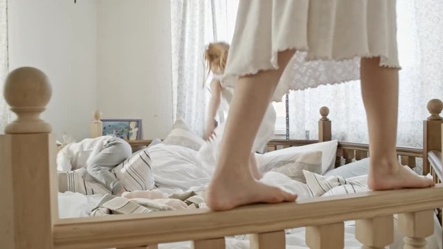 Slow motion tracking of legs of little girl in nightdress walking along side of big wooden bed while her younger sister and brother jumping and throwing pillows