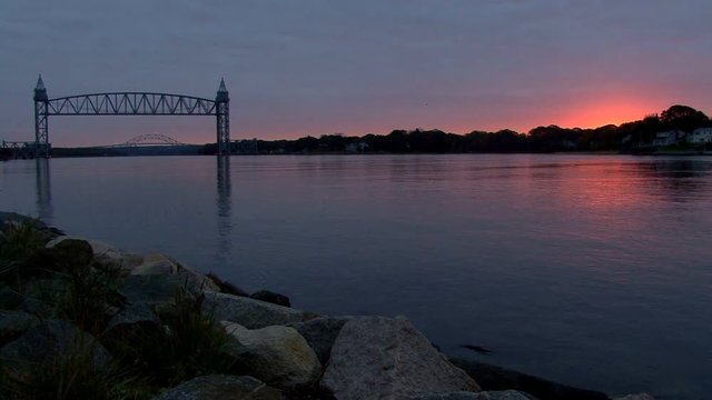Train bridge over tranquil waters of Cape Cod Canal frames distant Bourne Bridge in early morning .  Sunrises creating reddish pink colors in clouds
