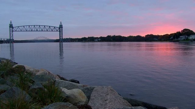 Train bridge over tranquil waters of Cape Cod Canal frames distant Bourne Bridge in early morning .  Sunrises creating reddish pink colors in clouds