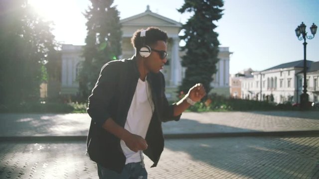 Young stylish hipster black man in white headphones and sun glasses dancing outdoor in city centre, having some fun