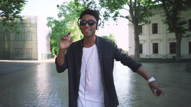 Young stylish hipster black man in white headphones and sun glasses dancing outdoor in city centre, having some fun