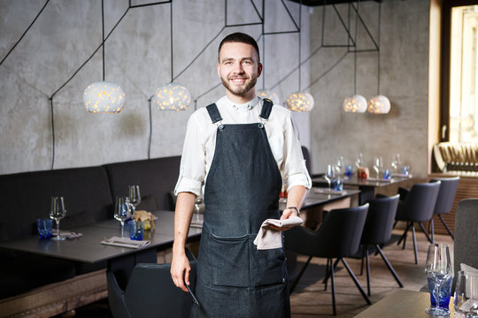A young, smiling waiter in a restaurant, standing next to the tables with a glass of wine. Dressed in an apron, will take an order holding a notebook and pen
