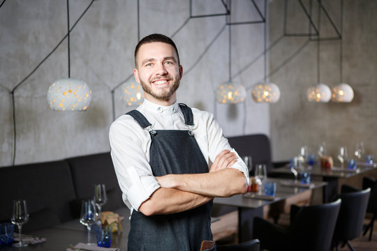 A young, smiling waiter in a restaurant, standing next to the tables with a glass of wine. He wears an apron, looks confidently, folded arms over his chest