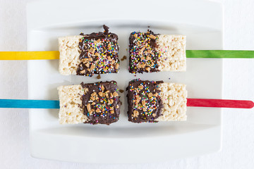 horizontal image of four rice krispie cakes on different coloured sticks dipped in chocolate and sprinkles on a white plate.