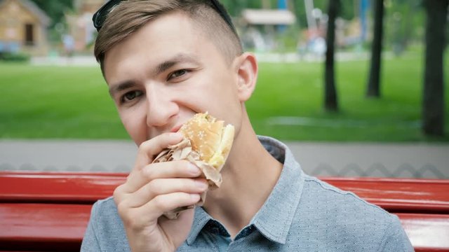 A man with a delight eating a hamburger sitting on a bench in a city park