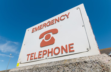 An retro old weathered and distressed emergency telephone sign, on a rustic seaside wall near the ocean and swimmers. The sign is old and vintage.