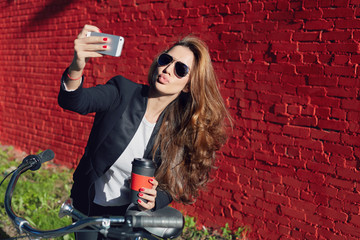 Fototapeta na wymiar Stylish bright portrait of attractive fashionable young beautiful Caucasian lady wearing formal suit, sunglasses riding bicycle to office making selfie during coffee break. Red brick wall background.