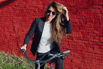 Fototapeta na wymiar Stylish bright portrait of attractive fashionable young beautiful Caucasian woman wearing formal suit and sunglasses, riding bicycle to office, looking at camera, posing on red brick wall background.