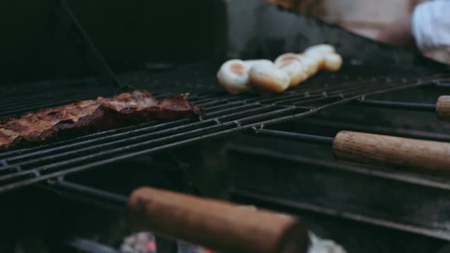 Close focus on big BBQ grill with pieces of smoked pork meat on it. Chief picks up grilled buns for small burges on background, unfocused. Private party catering concept