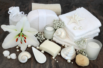 White lily flower with cleansing beauty treatment accessories including natural loofah, moisturising cream, soap, flannels, salt, natural sponge with shells and pearls.
