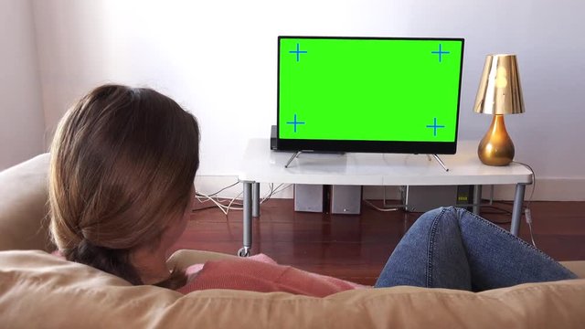 Girl Comfortably Lying On Couch Watching TV, Zoom In. Young woman watching television zoom in green screen. Shot behind models shoulders