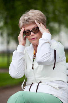 Elder European woman with tension-type headache resting on bench after running in park, outdoor