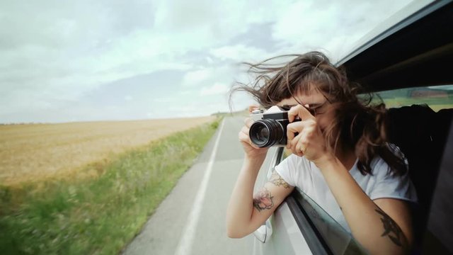 happy and excited young woman hangs out of car window, her brown hair blows in wind and she makes photos of landscapes and holiday spots on her vintage analog camera