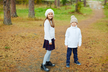 Fototapeta premium Brother and sister posing to camera, smiling outdoors in pine forest enjoying the walk on warm autumn day , both dressed in similar knit clothes