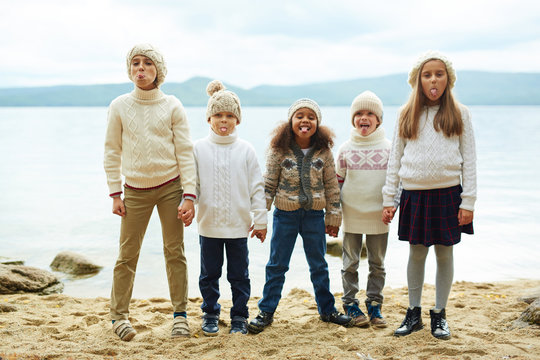 Group Of Cheerful Children Standing By Lake In Row, Sticking Out Tongues While Playing Outdoors On Warm Autumn Day All Dressed In Similar Knit Clothes