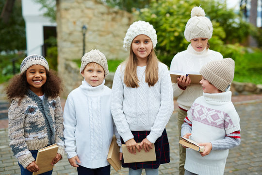 Children Of Various Ages Posing Outdoors Smiling To Camera And Holding Books, All Dressed In Similar Knit Clothes