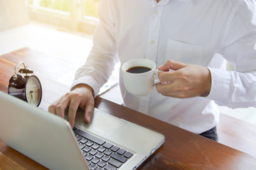 Selective focus of Young businessman using his laptop and drinking black coffee with clock and sunshine background.