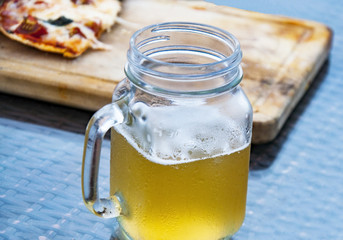 The misted glass of light beer and a piece of half-eaten pizza  on the table