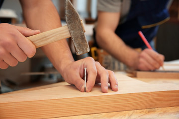 Carpenter driving nail into wooden board in workshop, closeup