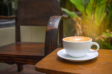 Hot coffee latte art heart shape in white cup on wooden table with empty wood chair and natura sunshinel background.