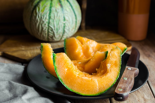 Whole and sliced ripe organic cantaloupe with juicy orange pulp, dark plate, knife, wood cutting board, rustic kitchen interior, cozy atmosphere