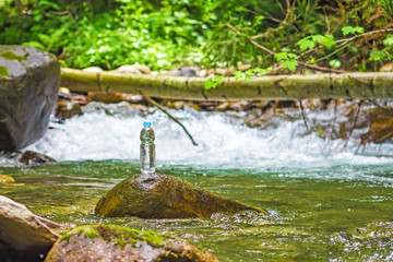 bottle of clear, clean, cold water stands on a stone in front of a waterfall. Photo for advertising of purified water and beverages