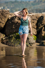 California girl walking along shiny, wet sand of waters edge with hand in hair looking down. 