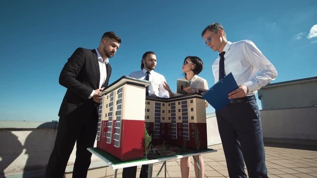 Group of engineers and architects in a meeting standing outdoors on a pation discussing a model of office or apartment blocks in a low angle view against blue sky.