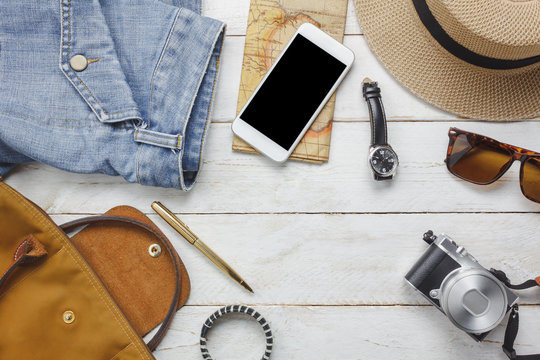 Top view accessoires to travel with women clothing concept. essential items are white mobile phone,watch,bag,hat,map,camera,necklace,trousers and sunglasses on white wood table.copy space.flat lay.