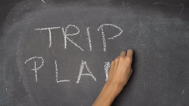 Woman's hand writing "TRIP PLAN" with white chalk on blackboard