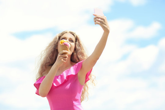 Beautiful young woman with ice cream taking selfie outdoors