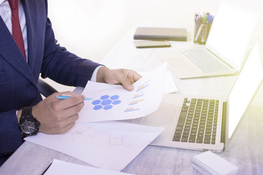 Close up view of a young businessman working on his desk white background. Two computers on the table. Man looking at paper charts and at laptop screens. Red tie and blue suit.