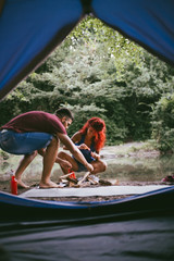 Obraz premium Happy young couple camping and hiking on wild mountain river. They trying to make campfire or bonfire and smiling. View from the tent. 