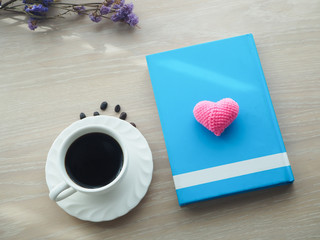 wood office table with hot black coffee cup, dried purple flower and pink heart shape sign on blank blue cover book.