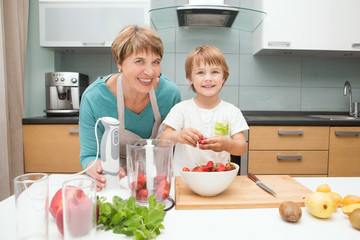 Happy grandmother and her little grandson making smoothie with fresh fruit and berries in the blender in kitchen. Little  boy preparing fruit cocktail or smoothie for breakfast. Healthy eating.