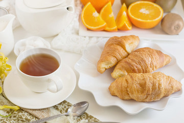 Fototapeta na wymiar Continental breakfast with gold french croissants fruits and cup of tea on white table in a Morning light. Breakfast concept