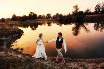 The bride and groom walk in the evening against the background of the lake in the red sunset. Overall plan