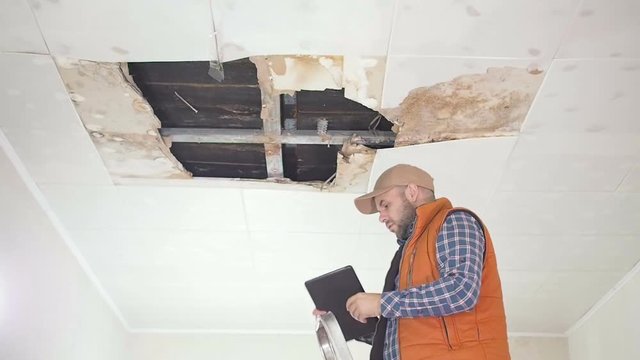 young man makes an emergency inspection of ceiling and use Tablet PC. public utilities. Ceiling panels damaged huge hole in roof from rainwater leakage.Water damaged ceiling , Insurance agent.