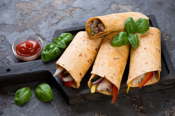 Tortilla rolls with roasted chicken meat and fresh vegetables on a black wooden serving tray, studio shot