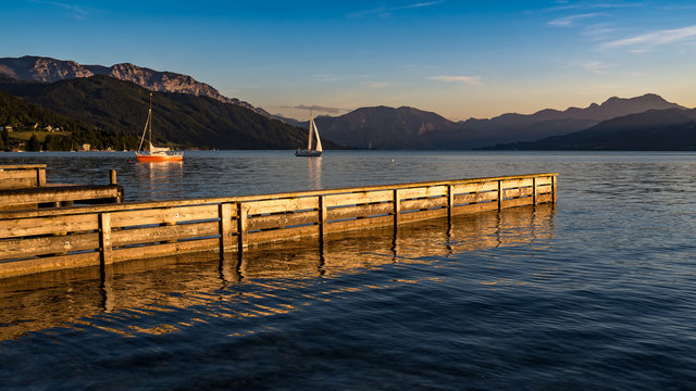 Old wooden pier in small rural town Steinbach am Attersee in Austria during the early sunset time sailboat is departing